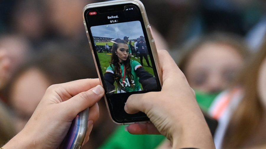 A young woman takes the first shot of her BeReal at a sporting event. The next shot will flip from the selfie camera to the back camera, taking a picture of her surroundings. 